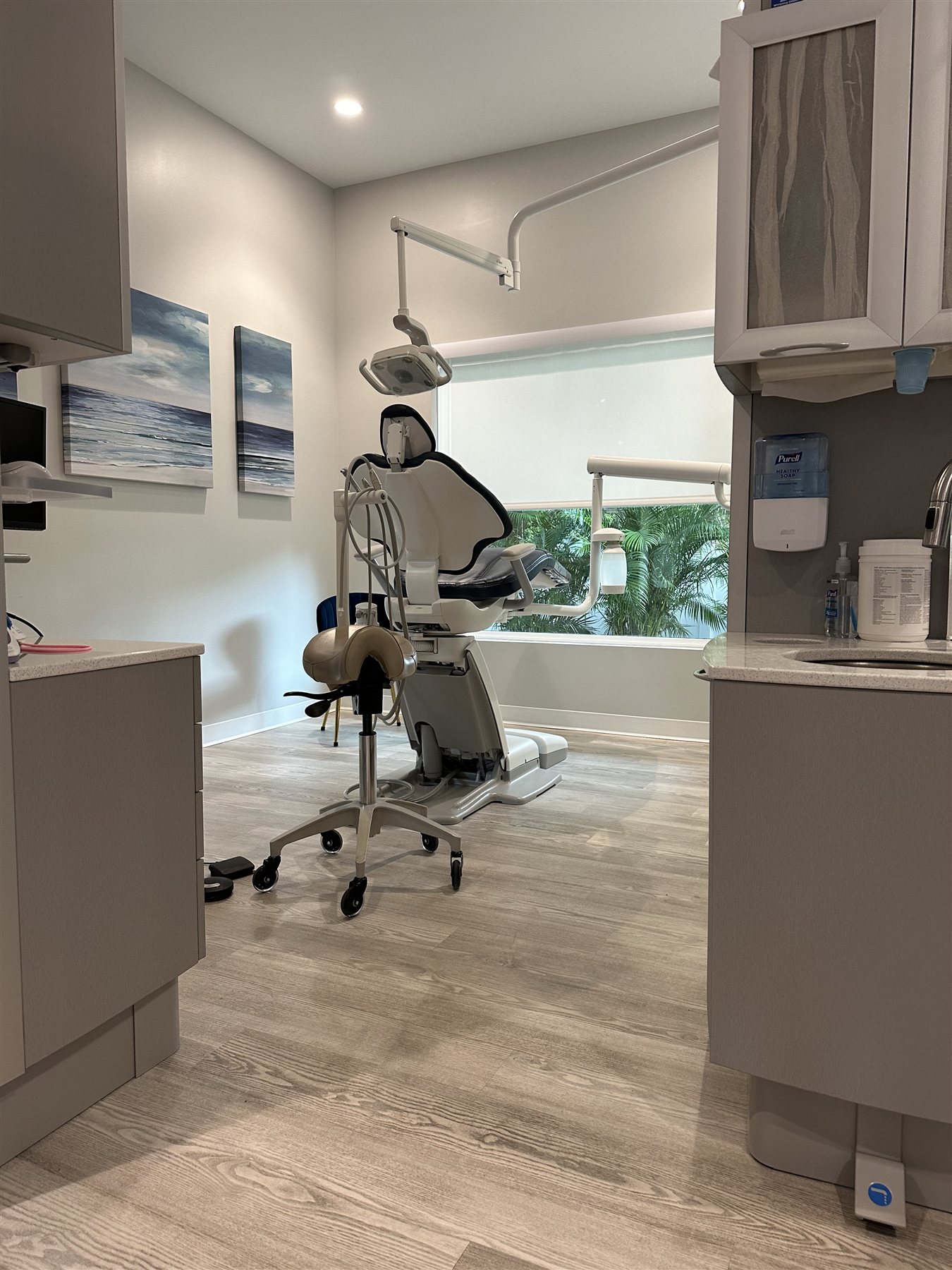 A dental treatment room with a chair beside a large window and coastal artwork.